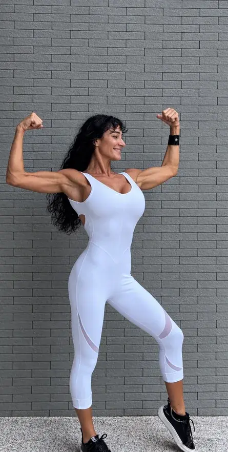 Anna smiling and flexing both arms in a white jumpsuit against a gray brick wall.