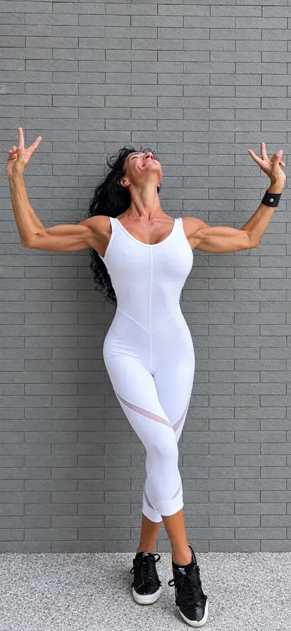 Anna in a white jumpsuit, arms raised high and smiling up, against a gray brick wall.